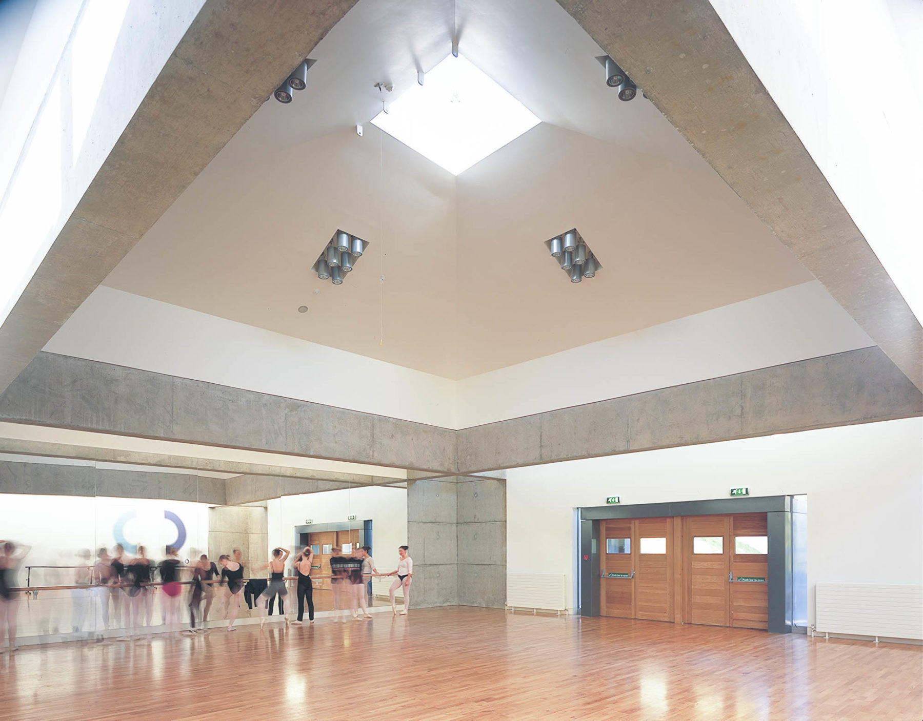 Large square studio at DanceBase Edinburgh, dancers training by mirror. Studio is lit from the skylight in the trapezoid roof. Malcolm Fraser Architects